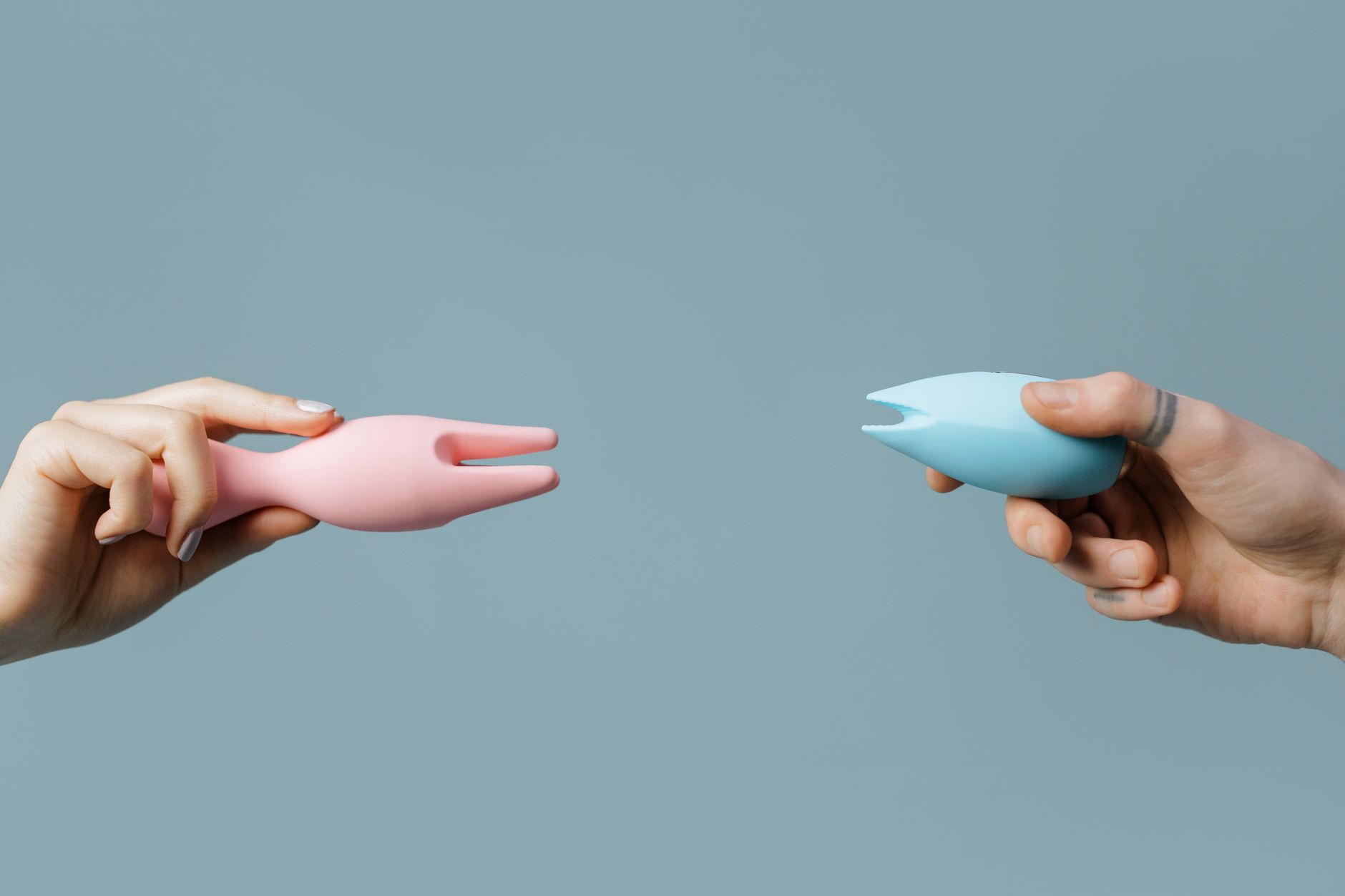 Hands holding silicone vibrators in pink and blue against a soft pastel background